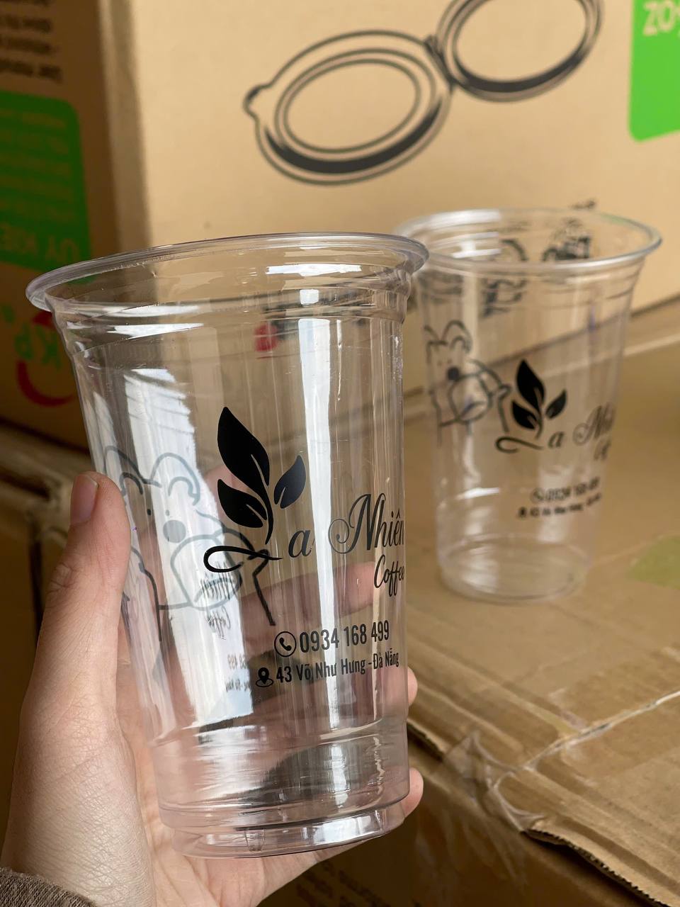 Ly nhựa UKP là gì? 5 lưu ý chọn chất liệu cho đồ uống 2026 5 Ly nhựa PET trong suốt in logo thương hiệu quán và thông tin liên hệ, được cầm tay với nền là hộp carton chứa sản phẩm tại Đà